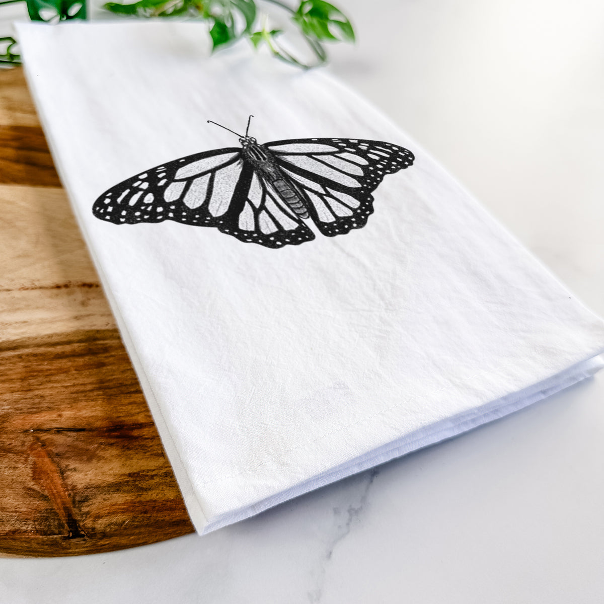 Close-up of a folded white tea towel on a wooden board, showing a detailed black ink illustration of a monarch butterfly with its wings spread.