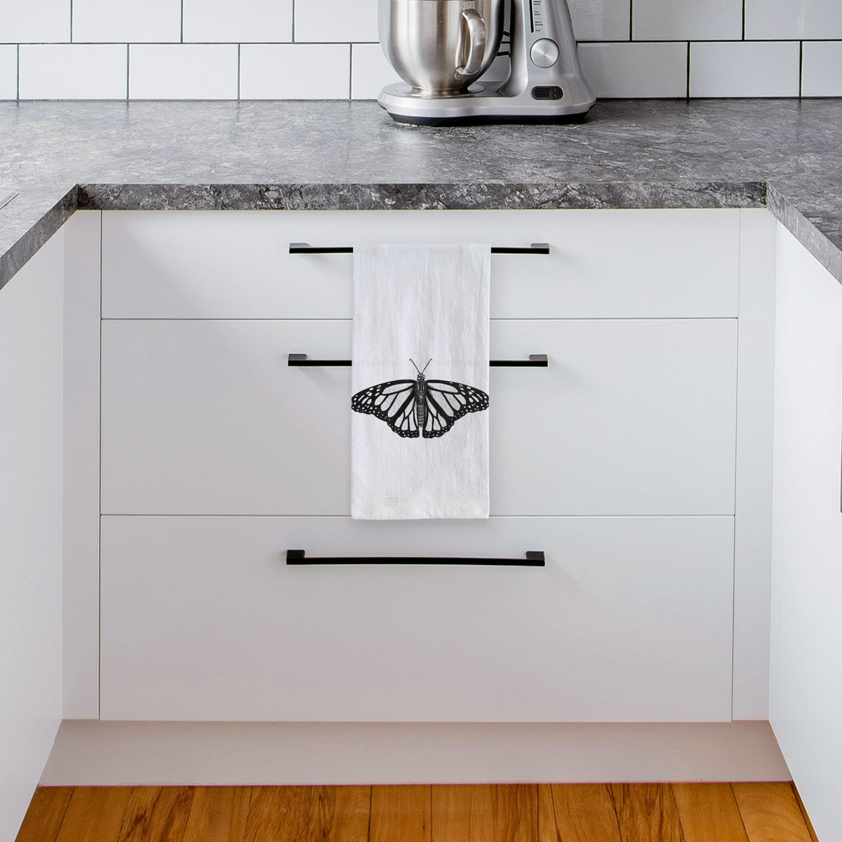 White tea towel printed with a detailed black ink illustration of a monarch butterfly with its wings spread, hanging from a drawer handle in a bright white kitchen.