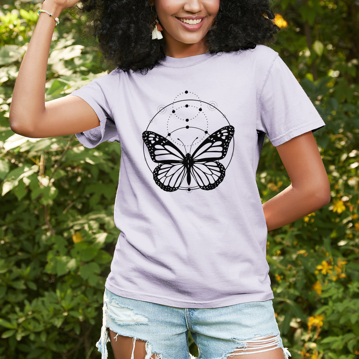 Woman wearing a Comfort Colors t-shirt printed with a black ink monarch butterfly inside a circle, beneath a line of moon phases, outdoors.