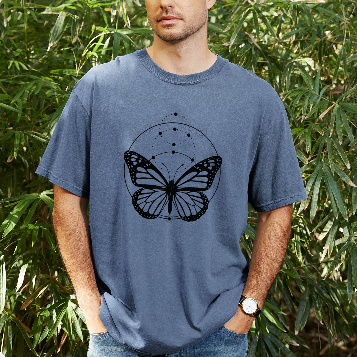 Man wearing a Comfort Colors t-shirt printed with a black ink monarch butterfly inside a circle, beneath a line of moon phases, outdoors.