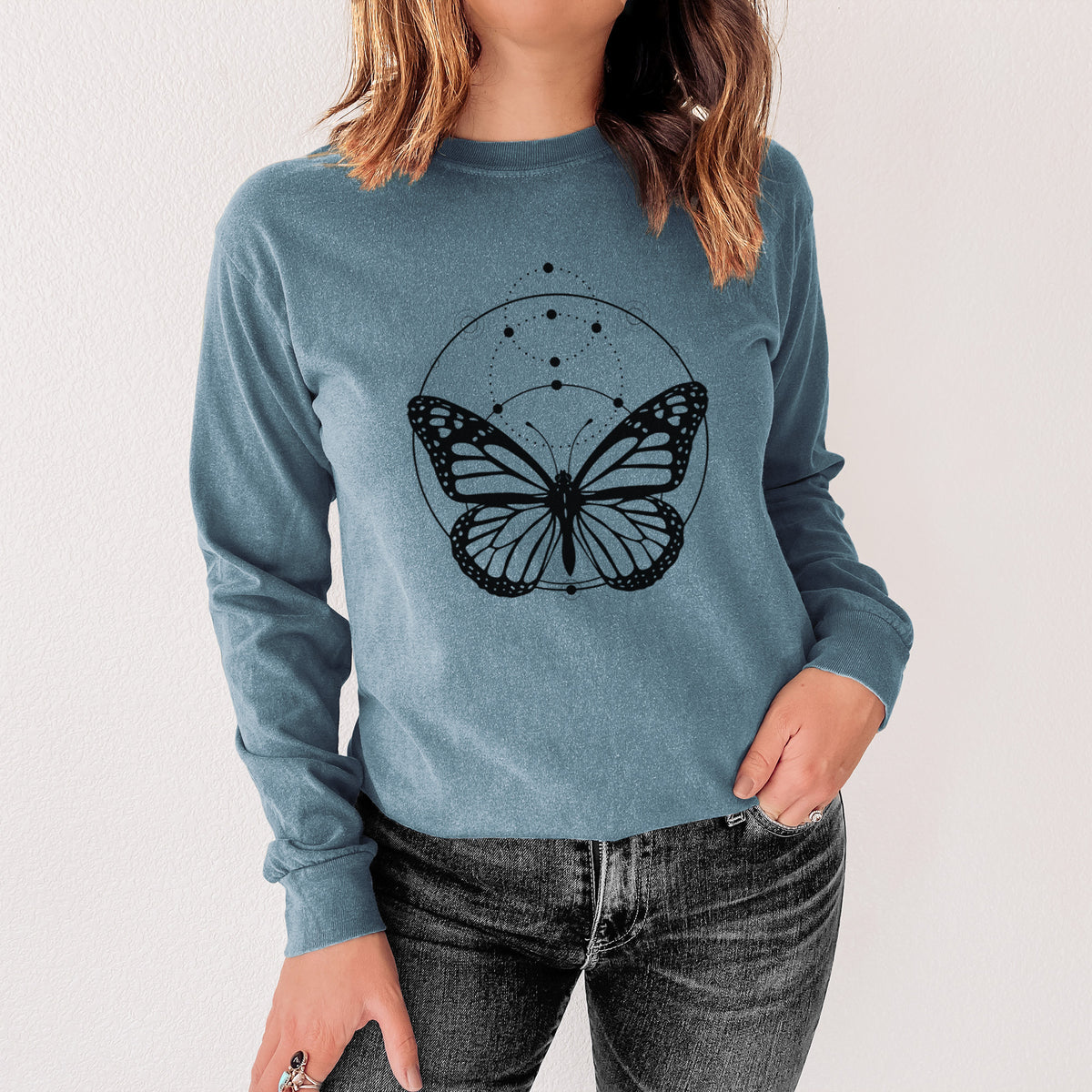 Steel blue Comfort Colors long sleeve shirt printed with a black ink monarch butterfly inside a circle, beneath a line of moon phases, shown in a lifestyle photo.