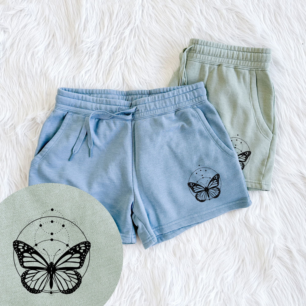 Two pairs of Cali Wave shorts in different colors printed with a black ink monarch butterfly inside a circle, beneath a line of moon phases, with a close-up of the print.