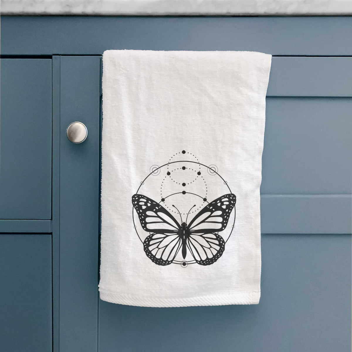 White hand towel printed with a black ink monarch butterfly inside a circle, beneath a line of moon phases, hanging on a cabinet door.