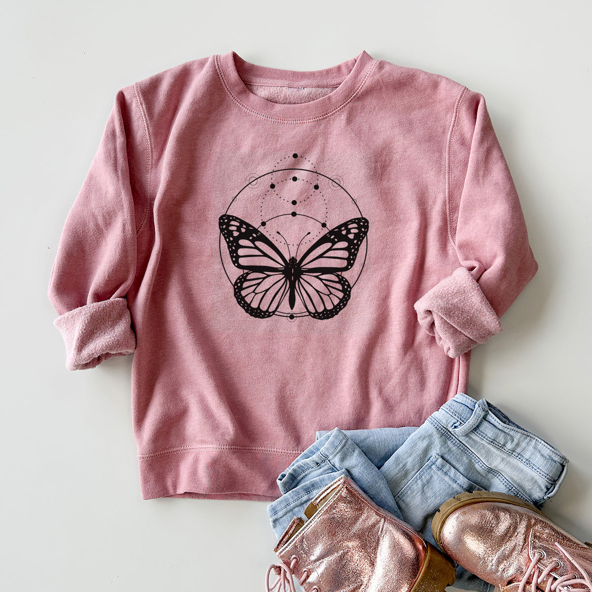 Youth crewneck sweatshirt printed with a black ink monarch butterfly inside a circle, beneath a line of moon phases, styled flat with jeans and boots.