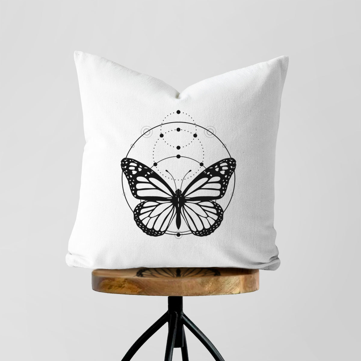 White pillow cover printed with a black ink monarch butterfly inside a circle, beneath a line of moon phases, on a wooden stool.