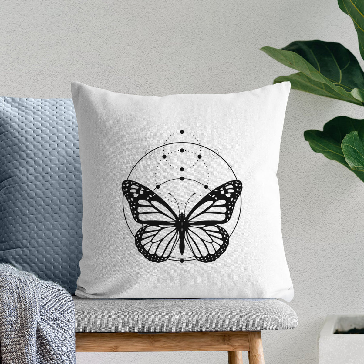 White pillow cover printed with a black ink monarch butterfly inside a circle, beneath a line of moon phases, on a bench beside plants.