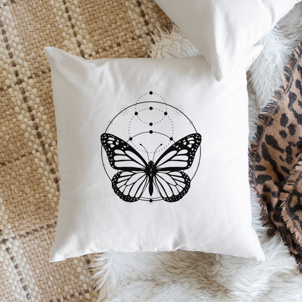 White pillow cover printed with a black ink monarch butterfly inside a circle, beneath a line of moon phases, on a woven chair.