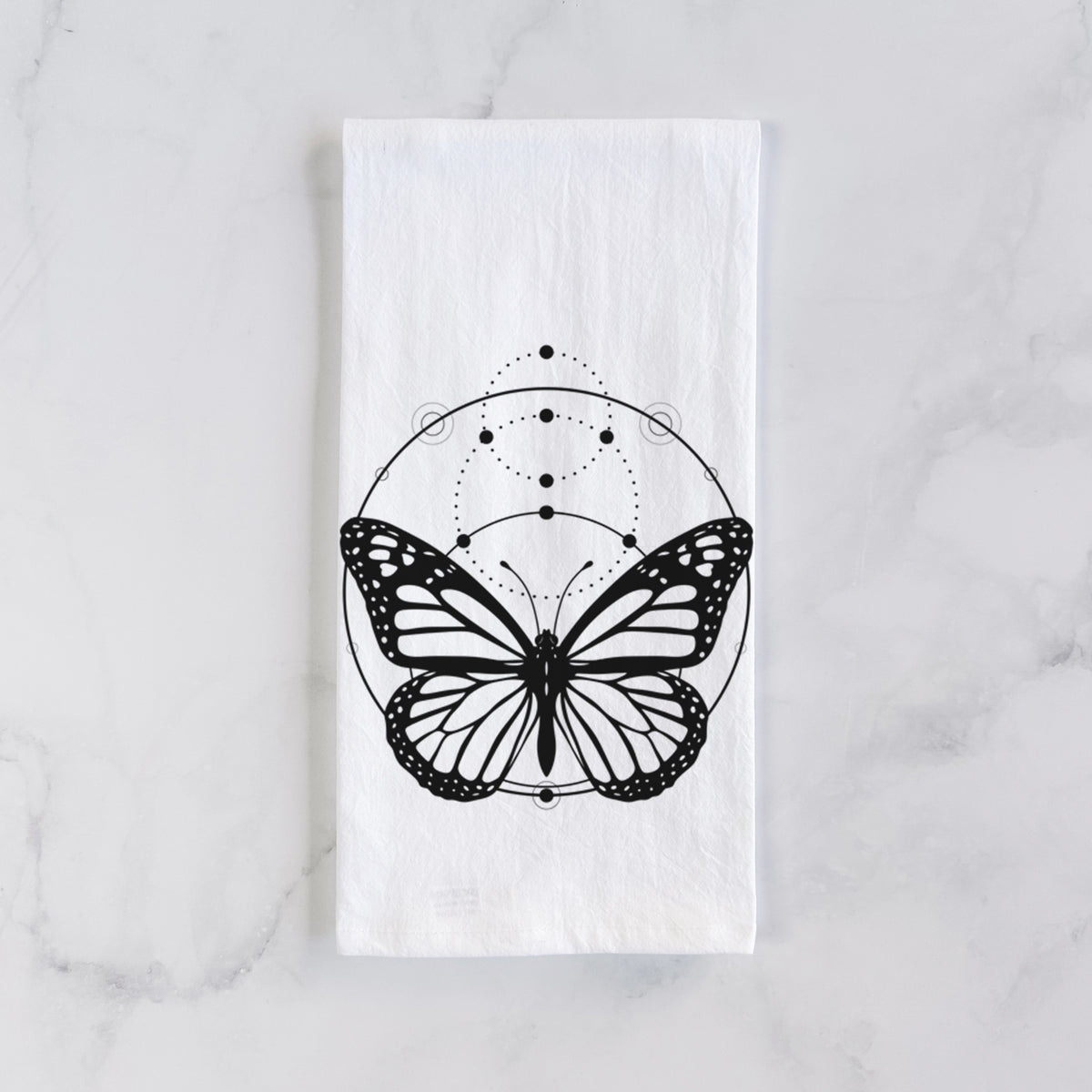 White cotton tea towel laid flat on marble, printed with a black ink monarch butterfly inside a circle, beneath a line of moon phases.