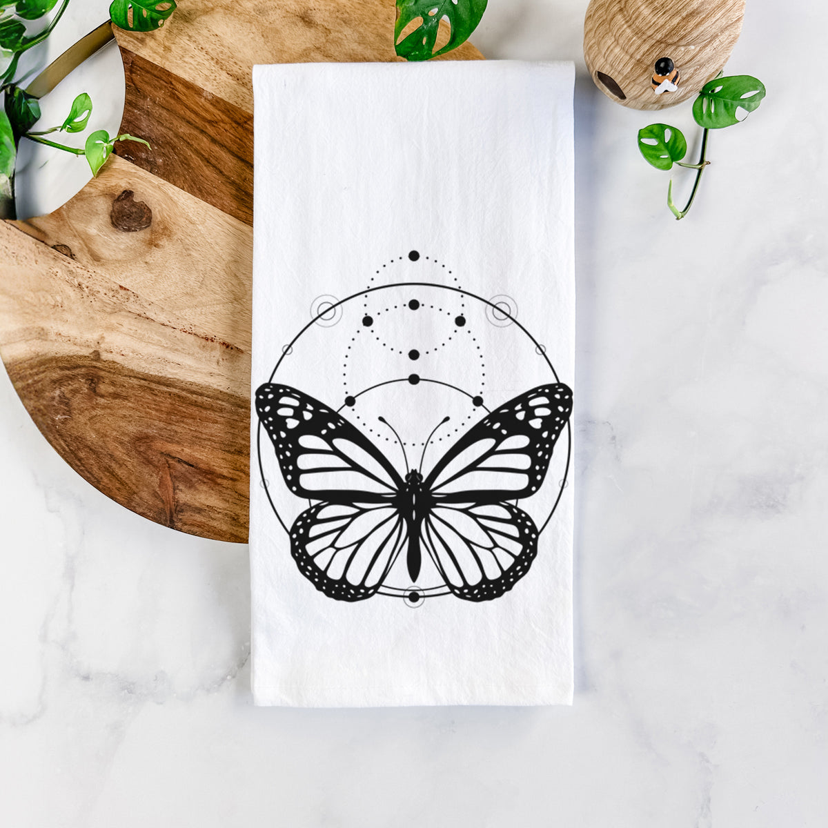 White cotton tea towel printed with a black ink monarch butterfly inside a circle, beneath a line of moon phases, on a marble counter beside a round wooden cutting board and greenery.
