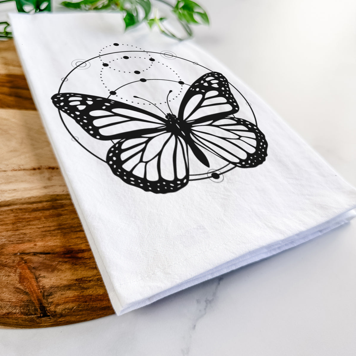 Close-up of a folded white tea towel on a wooden board, showing a black ink monarch butterfly inside a circle, beneath a line of moon phases.