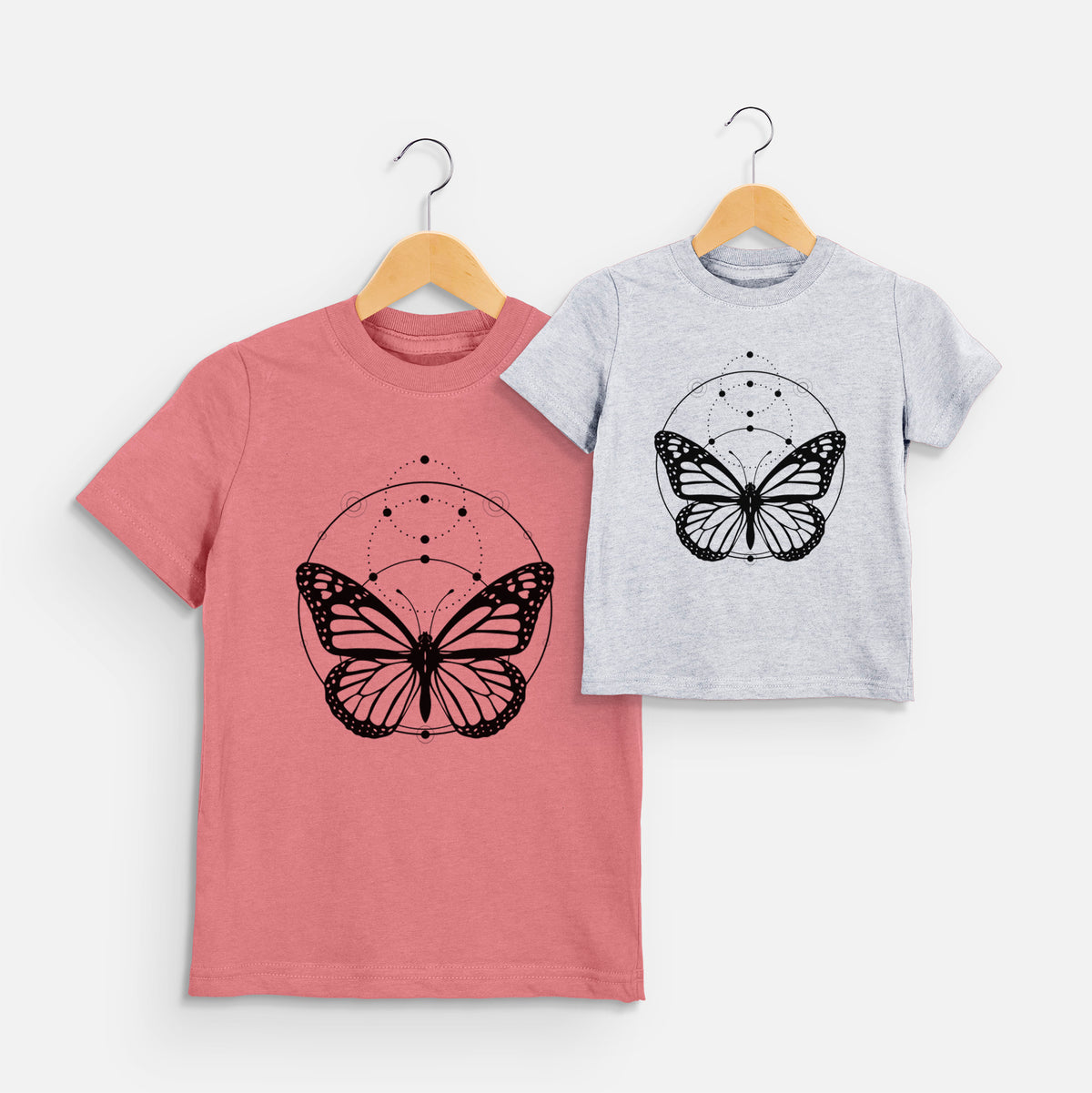 Two youth t-shirts in different colors on hangers, printed with a black ink monarch butterfly inside a circle, beneath a line of moon phases.