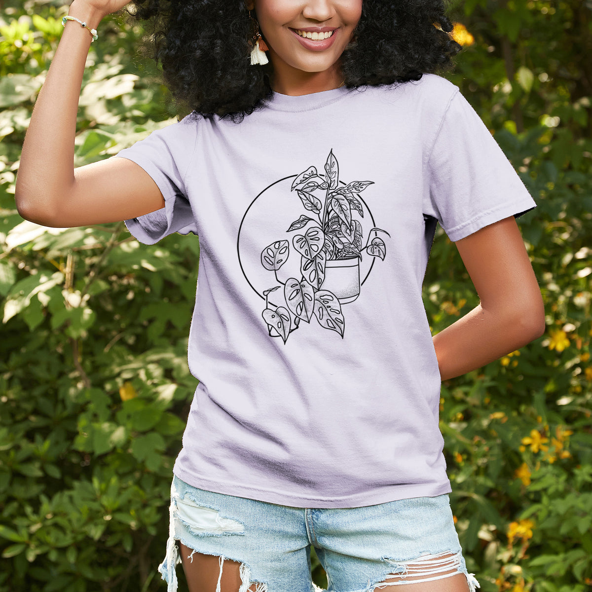 Woman wearing a Comfort Colors t-shirt printed with a black ink illustration of a potted Monsteradansonii plant with trailing vines in front of a circle, outdoors.