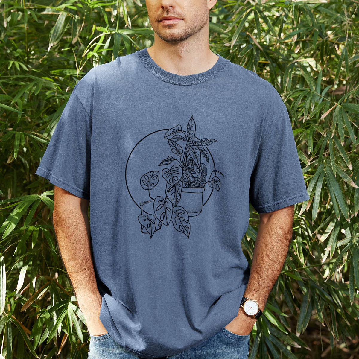 Man wearing a Comfort Colors t-shirt printed with a black ink illustration of a potted Monsteradansonii plant with trailing vines in front of a circle, outdoors.