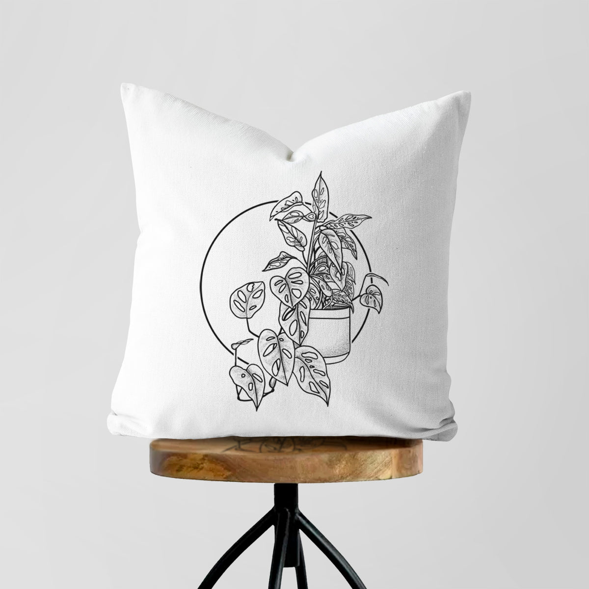 White pillow cover printed with a black ink illustration of a potted Monsteradansonii plant with trailing vines in front of a circle, on a wooden stool.