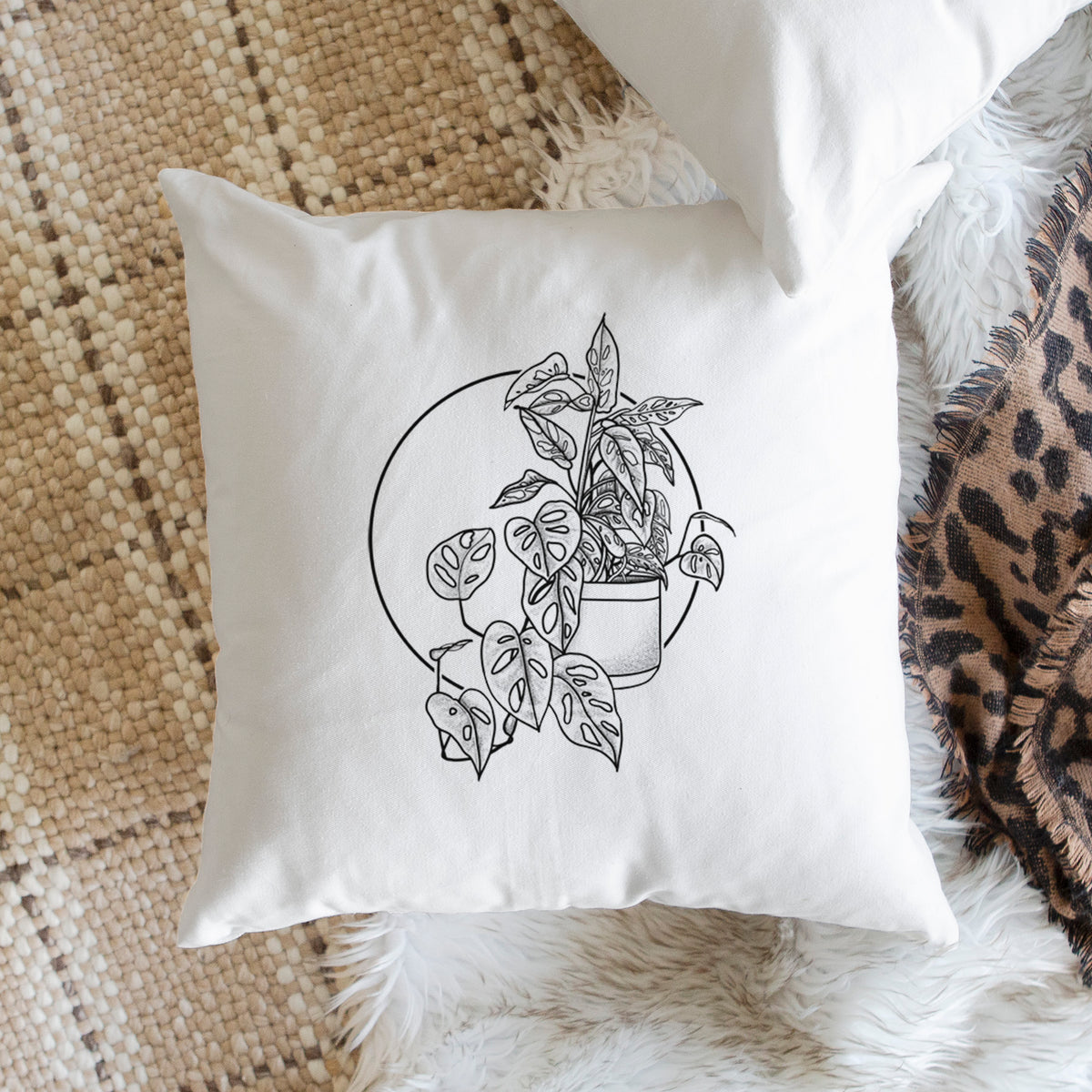 White pillow cover printed with a black ink illustration of a potted Monsteradansonii plant with trailing vines in front of a circle, on a woven chair.