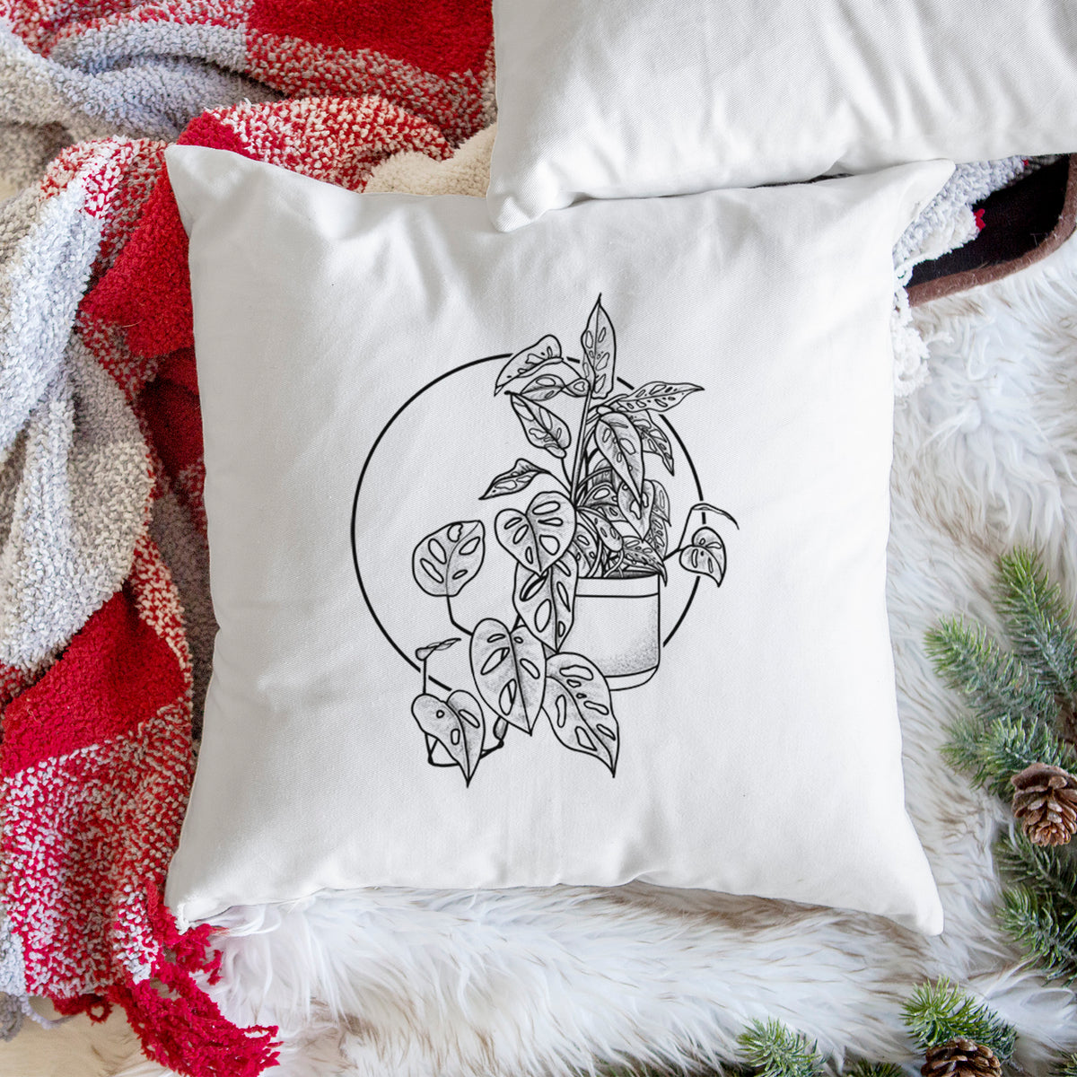 White pillow cover printed with a black ink illustration of a potted Monsteradansonii plant with trailing vines in front of a circle, on a woven chair.