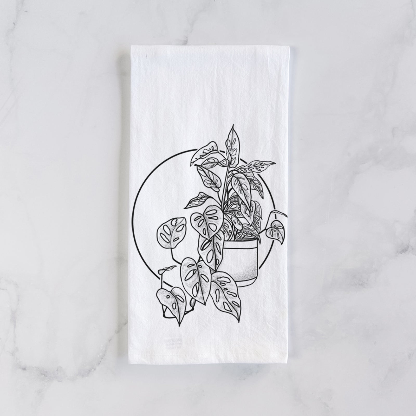 White cotton tea towel printed with a black ink illustration of a potted Monsteradansonii plant with trailing vines in front of a circle, on a marble counter beside a round wooden cutting board and greenery.