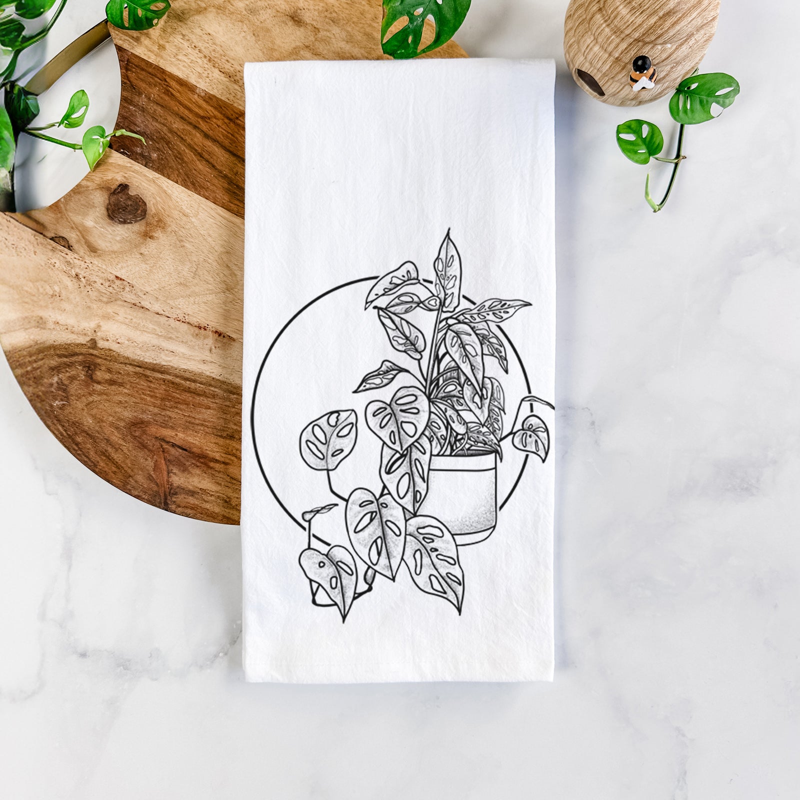 White cotton tea towel printed with a black ink illustration of a potted Monsteradansonii plant with trailing vines in front of a circle, on a marble counter beside a round wooden cutting board and greenery.