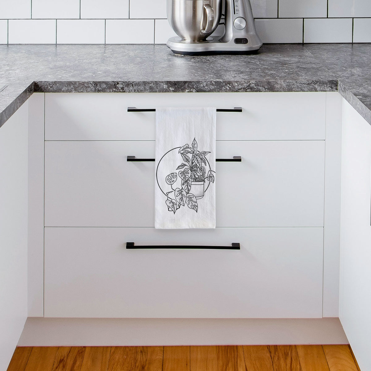 White tea towel printed with a black ink illustration of a potted Monsteradansonii plant with trailing vines in front of a circle, hanging from a drawer handle in a bright white kitchen.