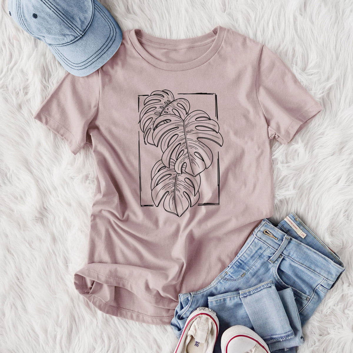 Women's relaxed fit cotton t-shirt printed with a black ink line drawing of Monstera deliciosa leaves inside a rectangle outline, styled flat on a white faux fur rug with jeans.