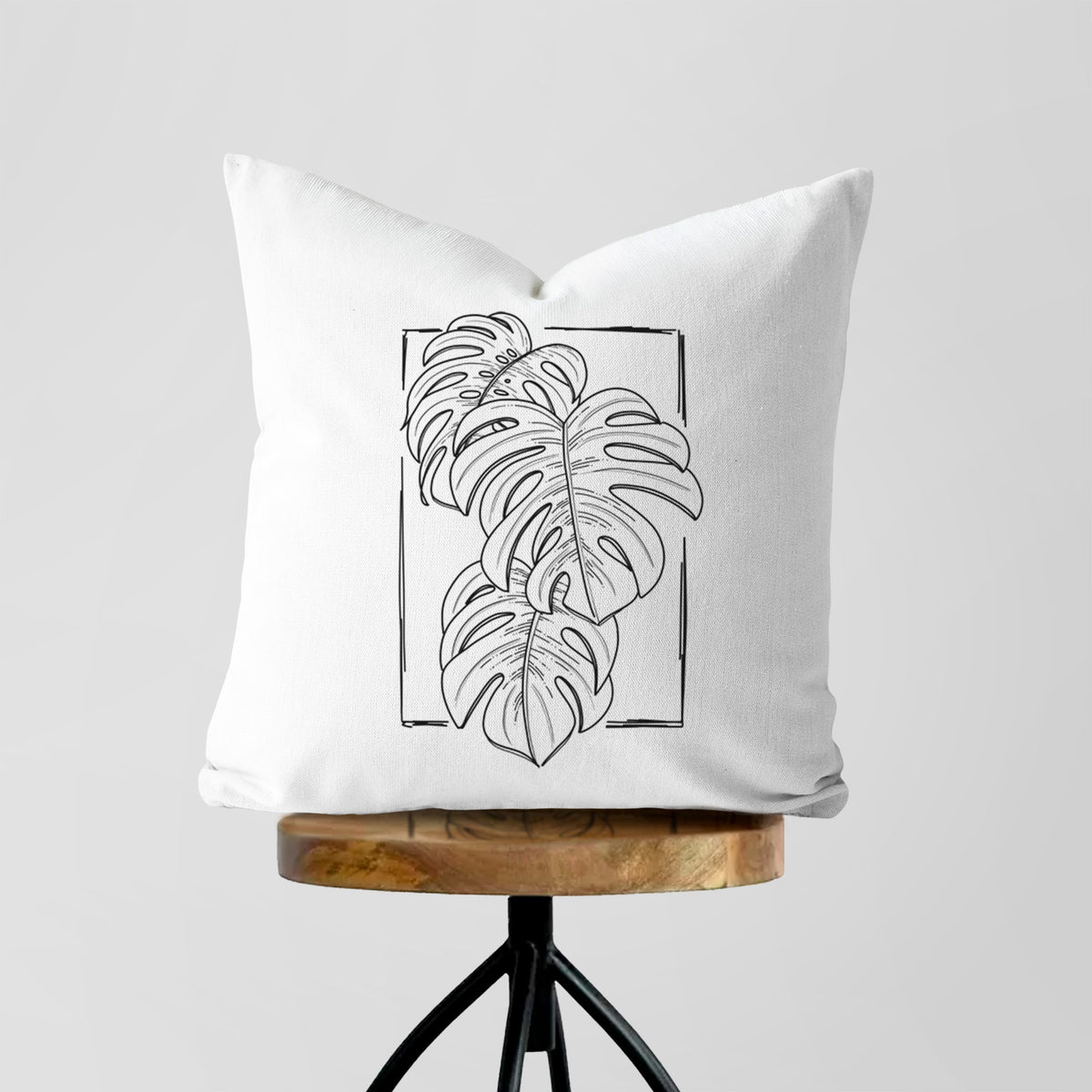 White pillow cover printed with a black ink line drawing of Monstera deliciosa leaves inside a rectangle outline, on a wooden stool.