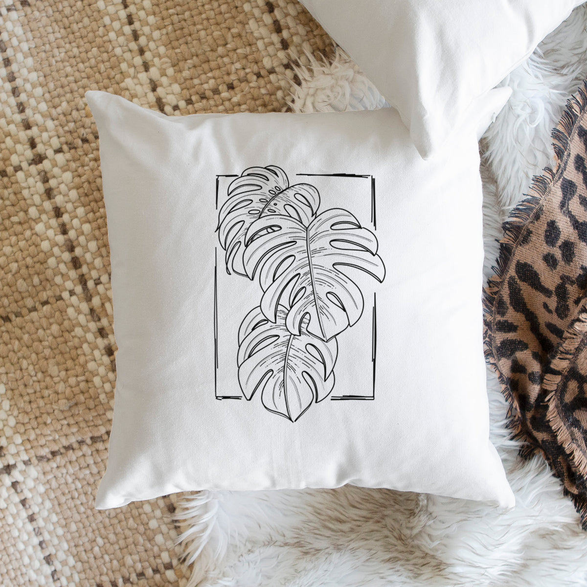 White pillow cover printed with a black ink line drawing of Monstera deliciosa leaves inside a rectangle outline, on a woven chair.