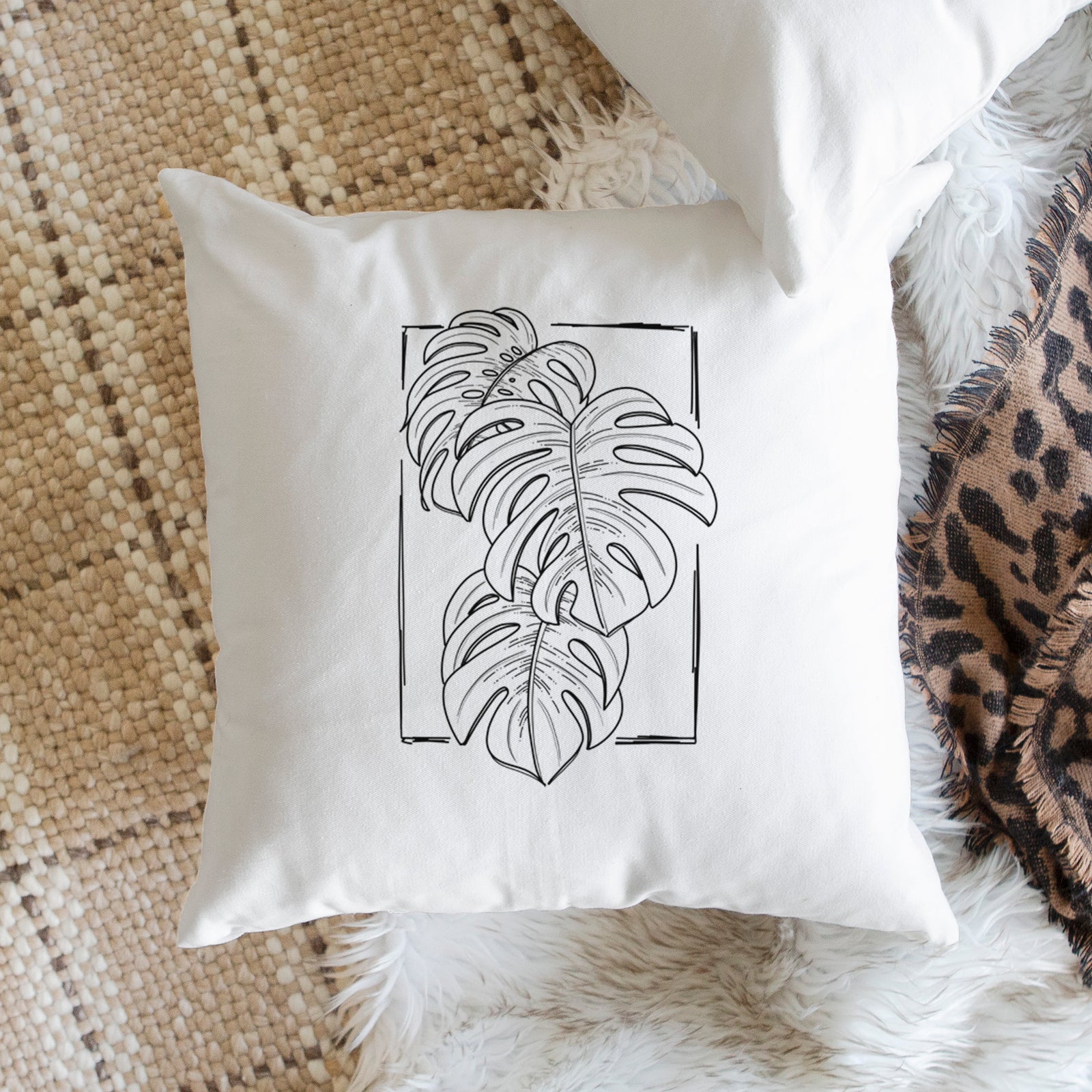 White pillow cover printed with a black ink line drawing of Monstera deliciosa leaves inside a rectangle outline, on a bench beside plants.