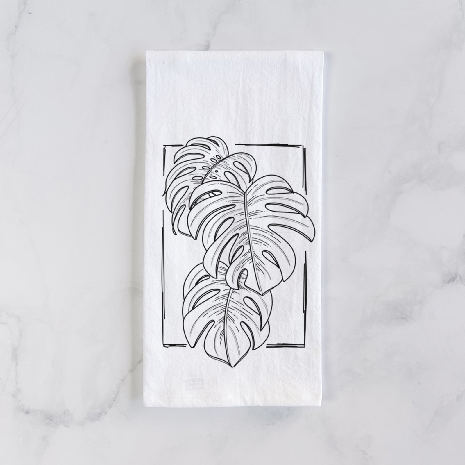 White cotton tea towel printed with a black ink line drawing of Monstera deliciosa leaves inside a rectangle outline, on a marble counter beside a round wooden cutting board and greenery.