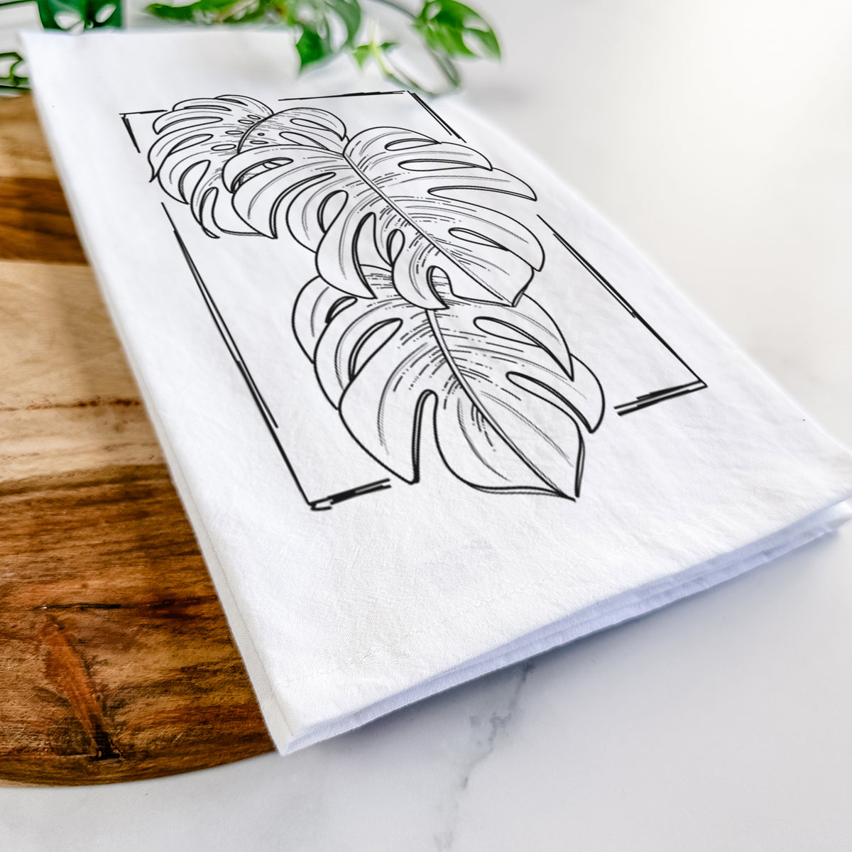 Close-up of a folded white tea towel on a wooden board, showing a black ink line drawing of Monstera deliciosa leaves inside a rectangle outline.
