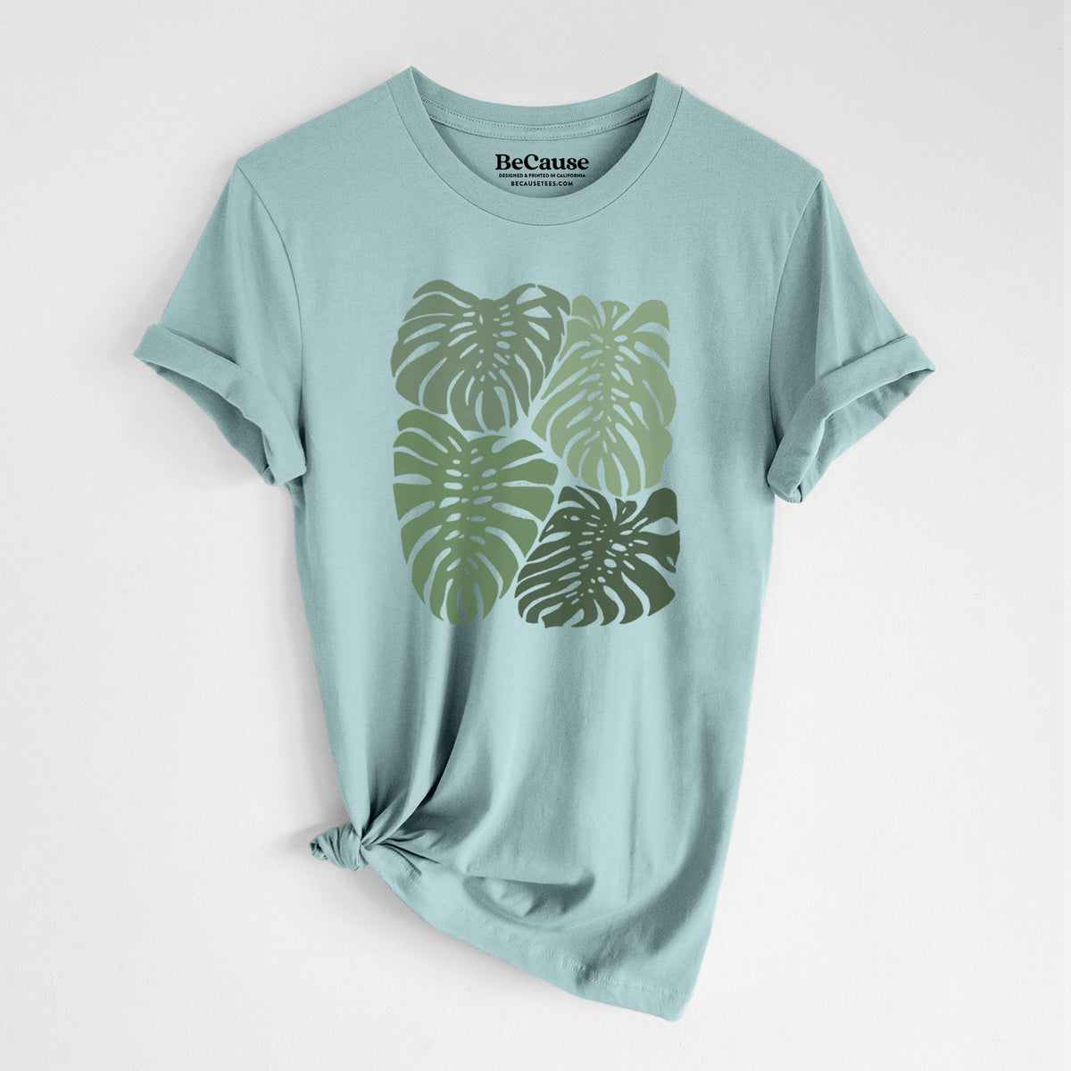 Dusty blue lightweight cotton t-shirt laid flat with the sleeves rolled, printed with a full-color illustration of large green Monstera leaves filling a square.