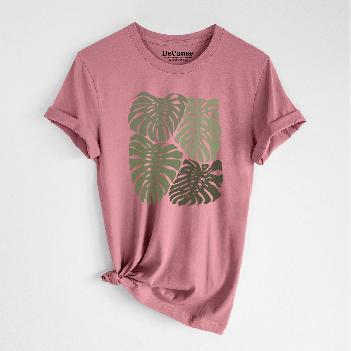 Mauve pink lightweight cotton t-shirt laid flat with the sleeves rolled, printed with a full-color illustration of large green Monstera leaves filling a square.