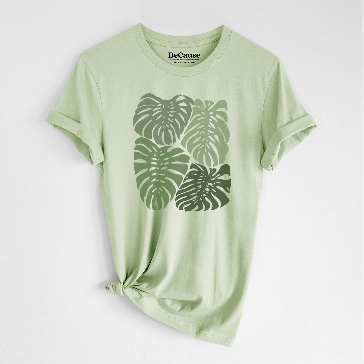 Spring green lightweight cotton t-shirt laid flat with the sleeves rolled, printed with a full-color illustration of large green Monstera leaves filling a square.