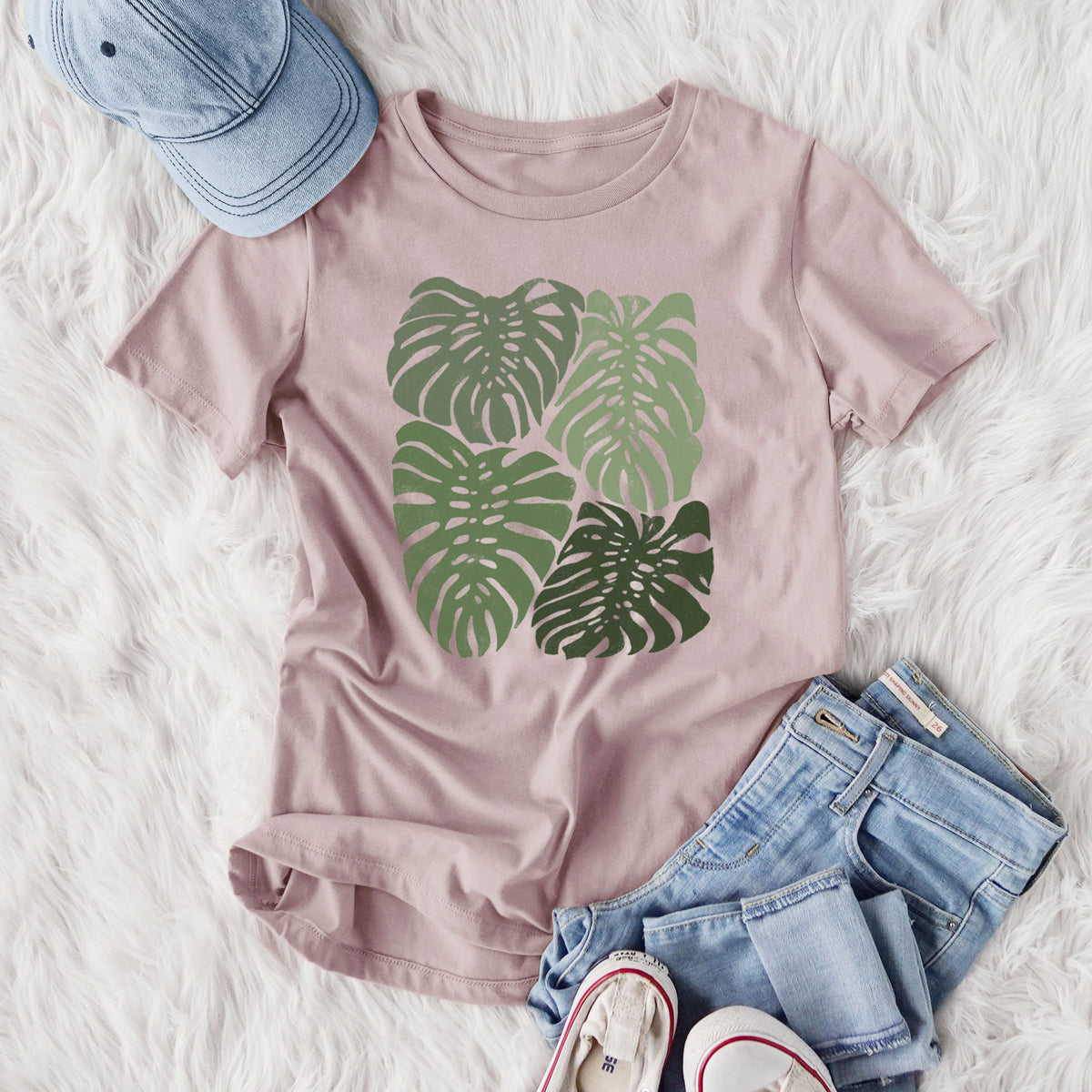 Women's relaxed fit cotton t-shirt printed with a full-color illustration of large green Monstera leaves filling a square, styled flat on a white faux fur rug with jeans.