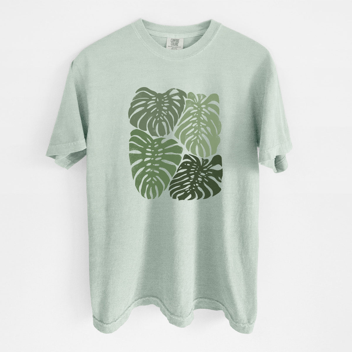 Bay green Comfort Colors t-shirt laid flat, printed with a full-color illustration of large green Monstera leaves filling a square.