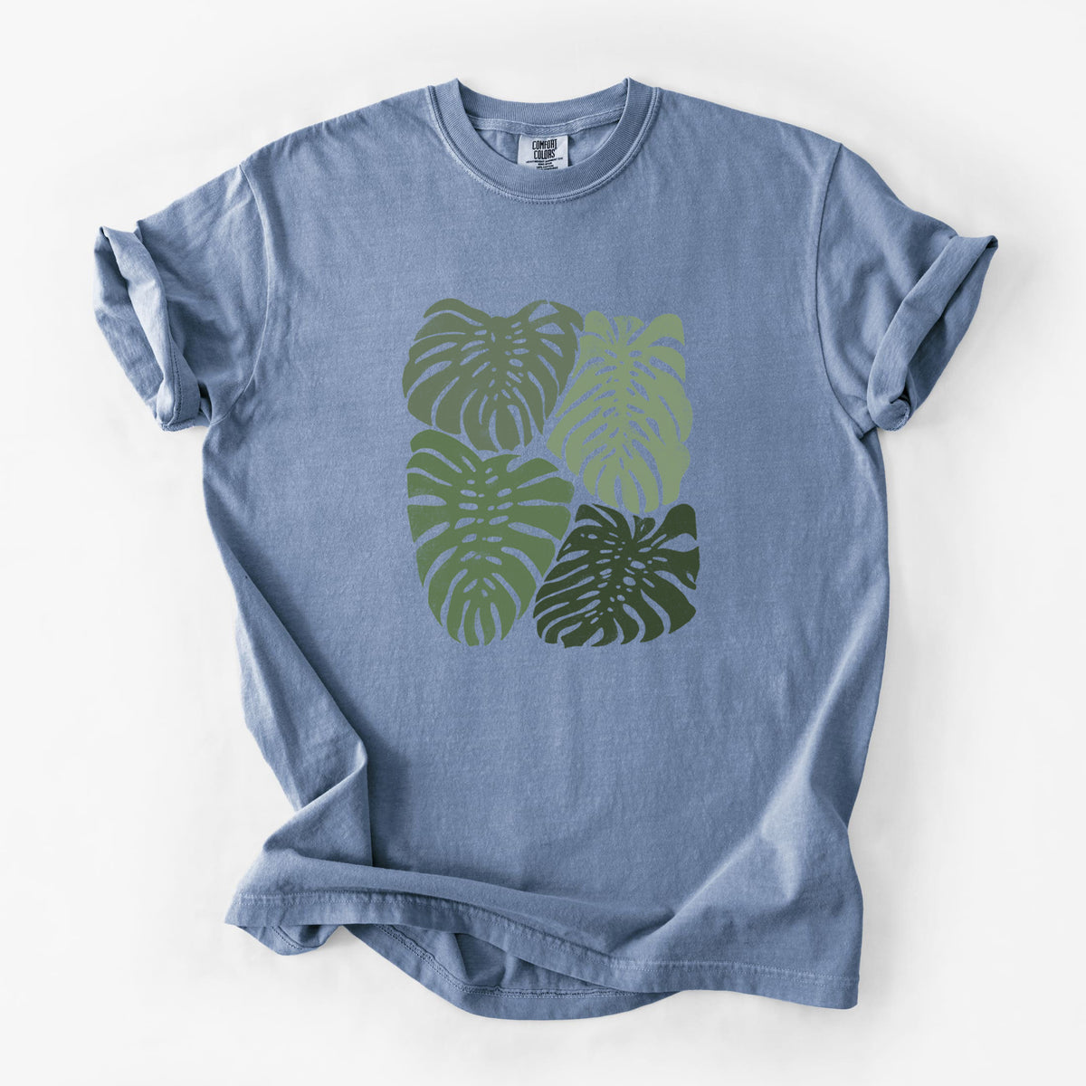 Blue jean Comfort Colors t-shirt laid flat, printed with a full-color illustration of large green Monstera leaves filling a square.