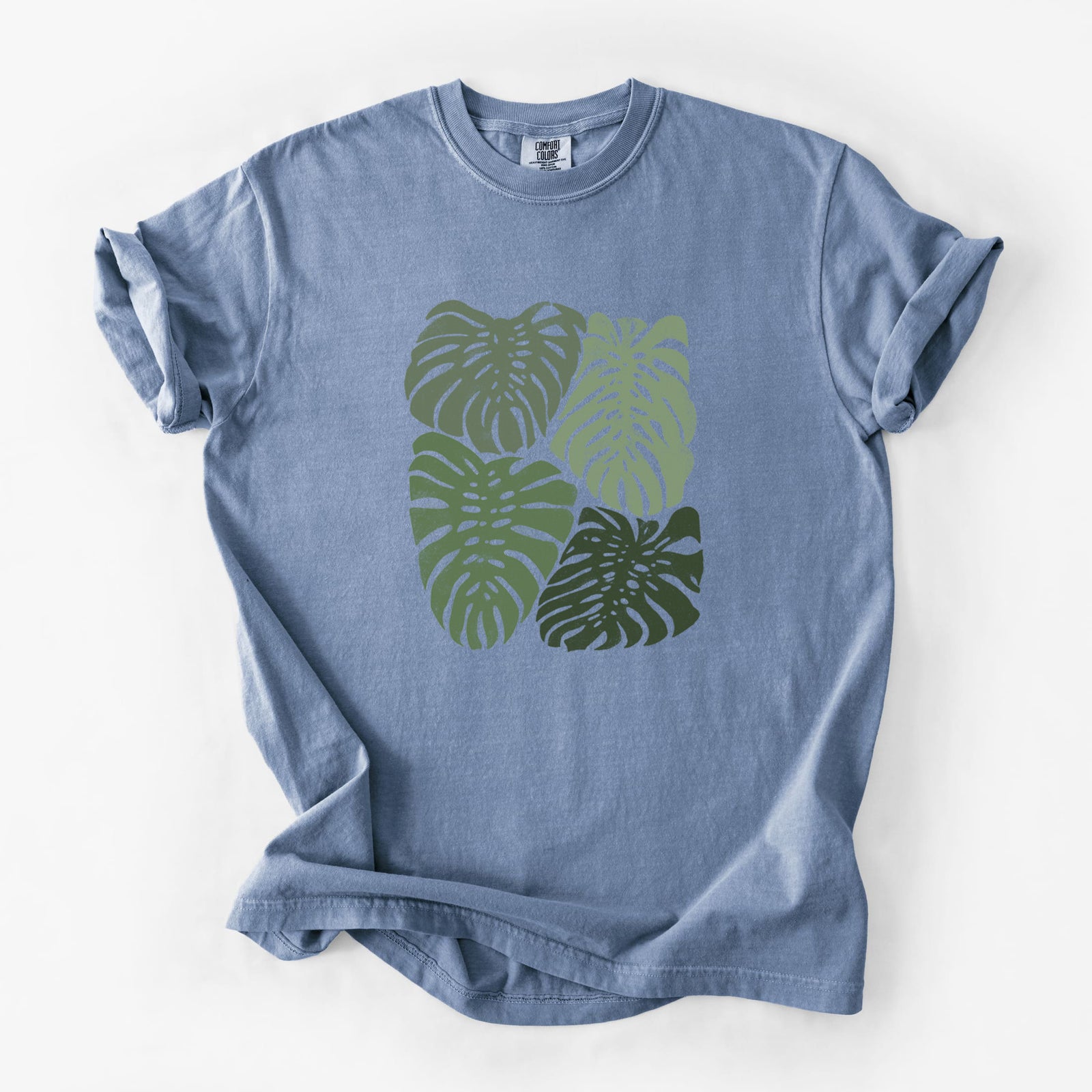 Blue jean Comfort Colors t-shirt laid flat, printed with a full-color illustration of large green Monstera leaves filling a square.