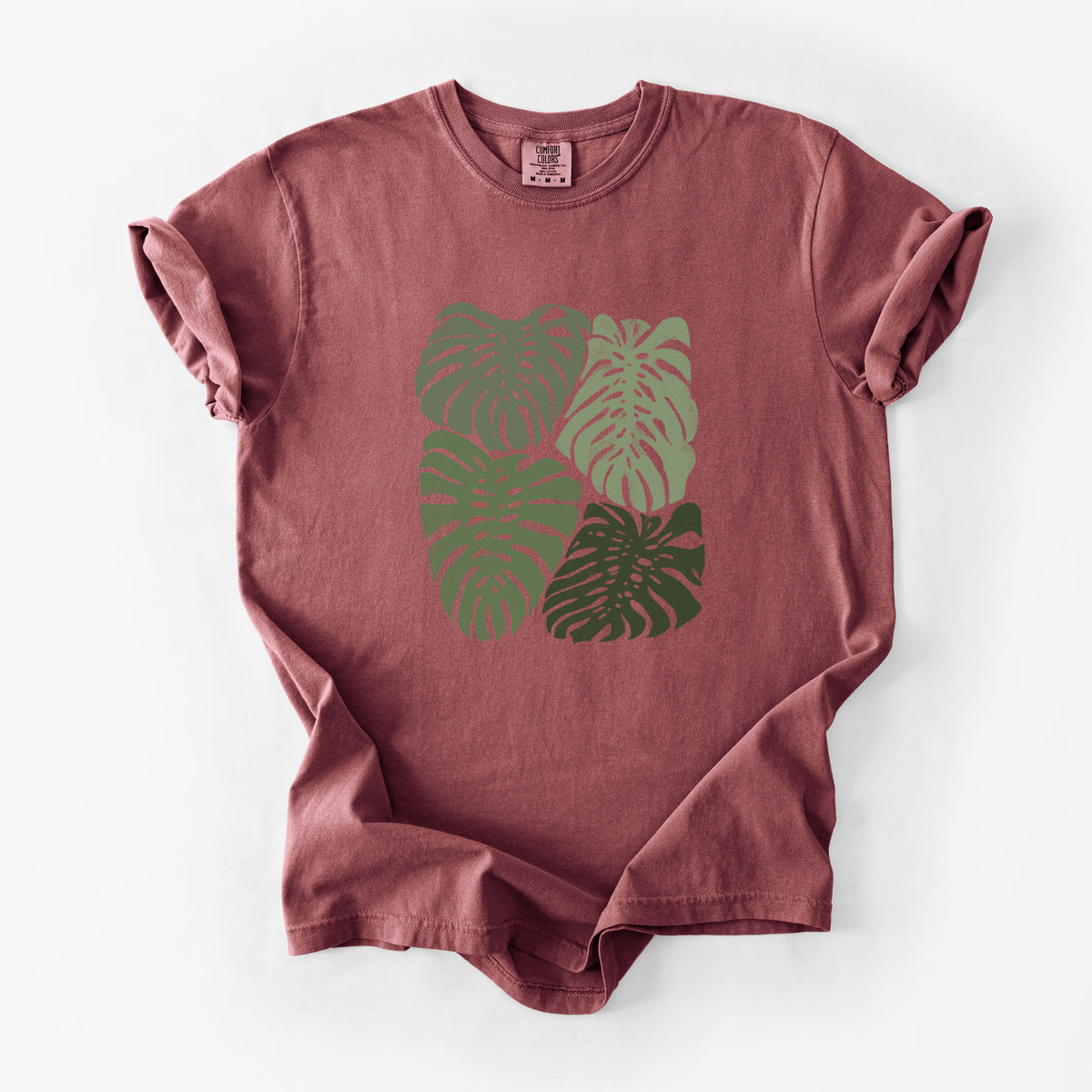 Brick red Comfort Colors t-shirt laid flat, printed with a full-color illustration of large green Monstera leaves filling a square.