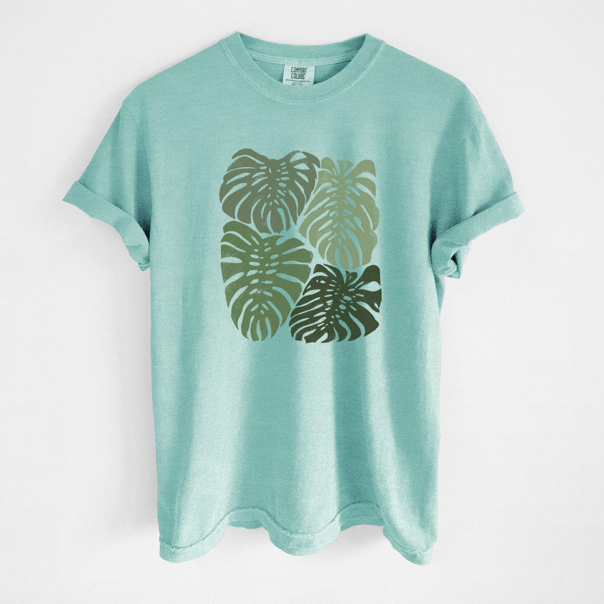 Chalky mint Comfort Colors t-shirt laid flat, printed with a full-color illustration of large green Monstera leaves filling a square.