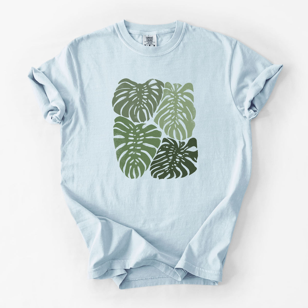 Chambray blue Comfort Colors t-shirt laid flat, printed with a full-color illustration of large green Monstera leaves filling a square.