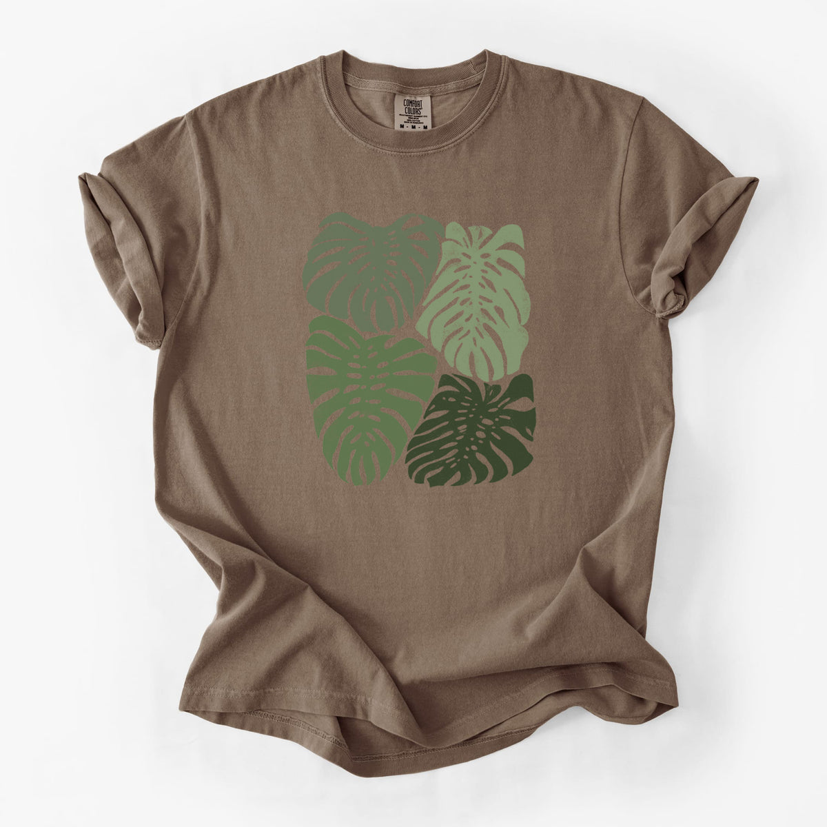 Espresso brown Comfort Colors t-shirt laid flat, printed with a full-color illustration of large green Monstera leaves filling a square.