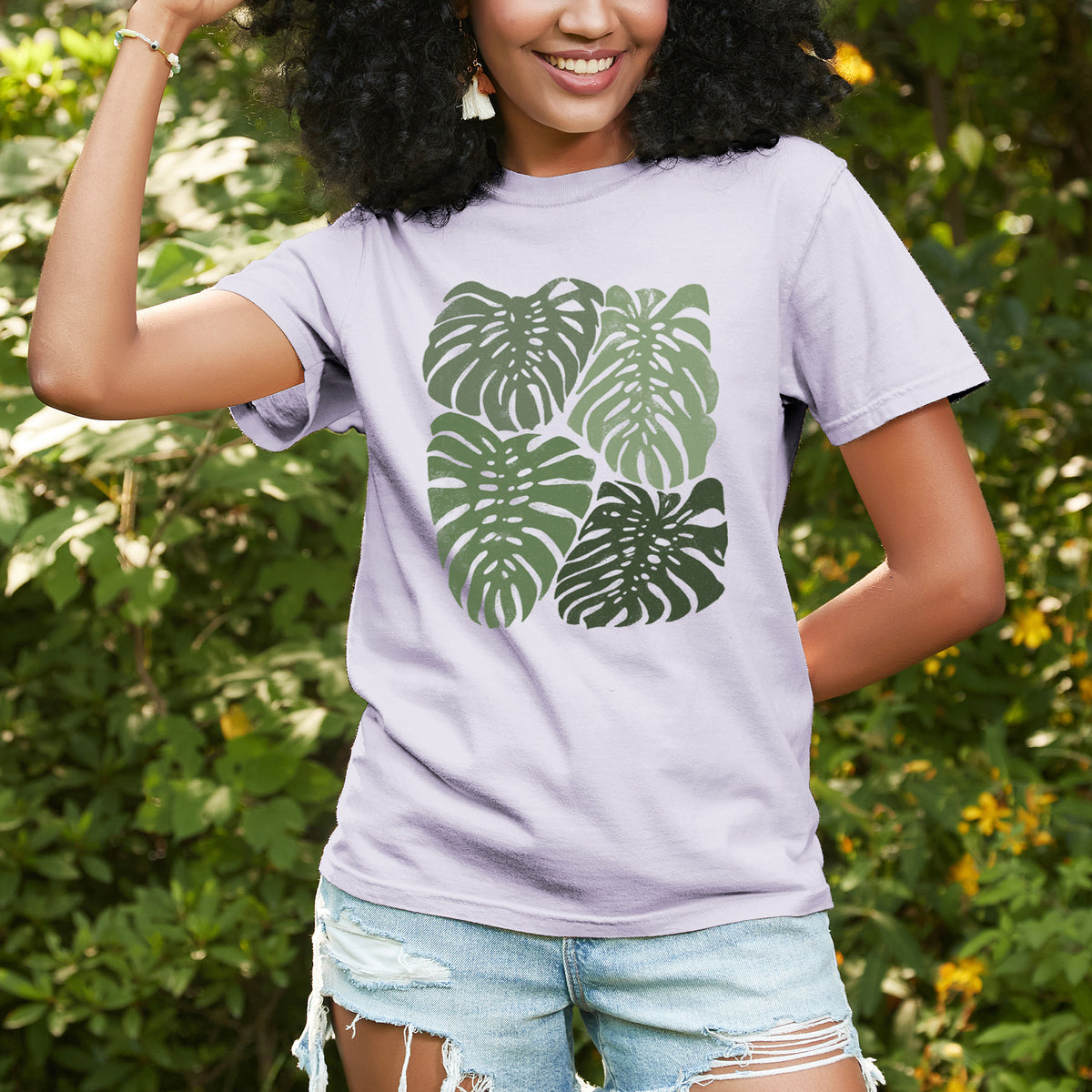 Woman wearing a Comfort Colors t-shirt printed with a full-color illustration of large green Monstera leaves filling a square, outdoors.