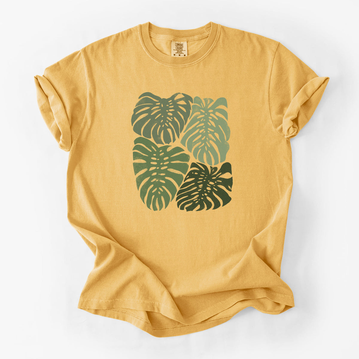 Golden wheat yellow Comfort Colors t-shirt laid flat, printed with a full-color illustration of large green Monstera leaves filling a square.