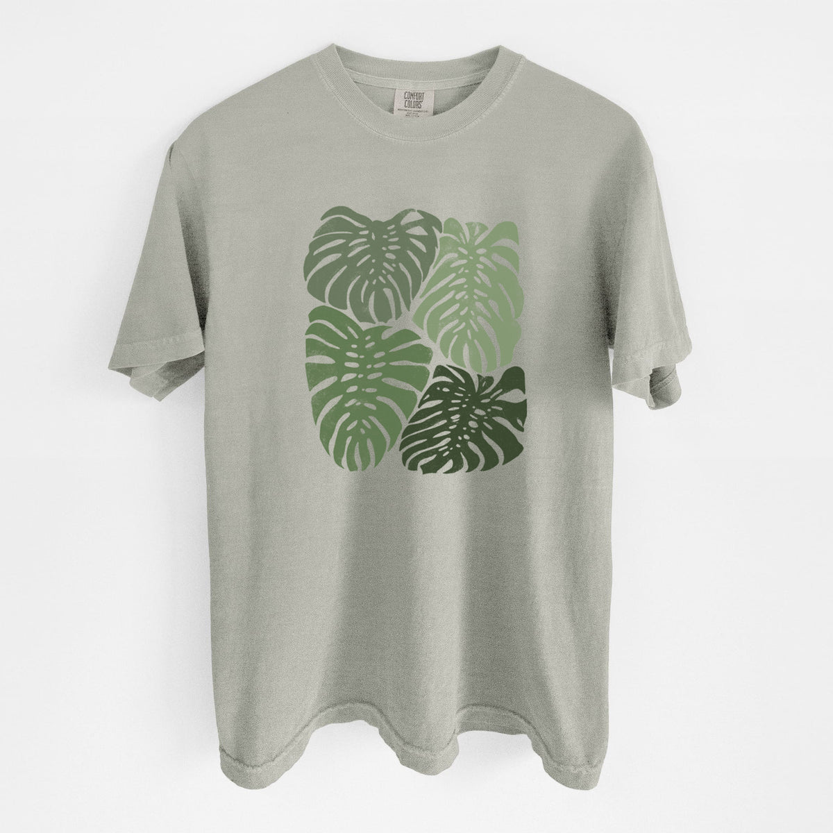 Sandstone beige Comfort Colors t-shirt laid flat, printed with a full-color illustration of large green Monstera leaves filling a square.