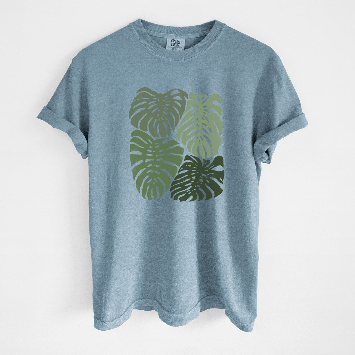 Steel blue Comfort Colors t-shirt laid flat, printed with a full-color illustration of large green Monstera leaves filling a square.