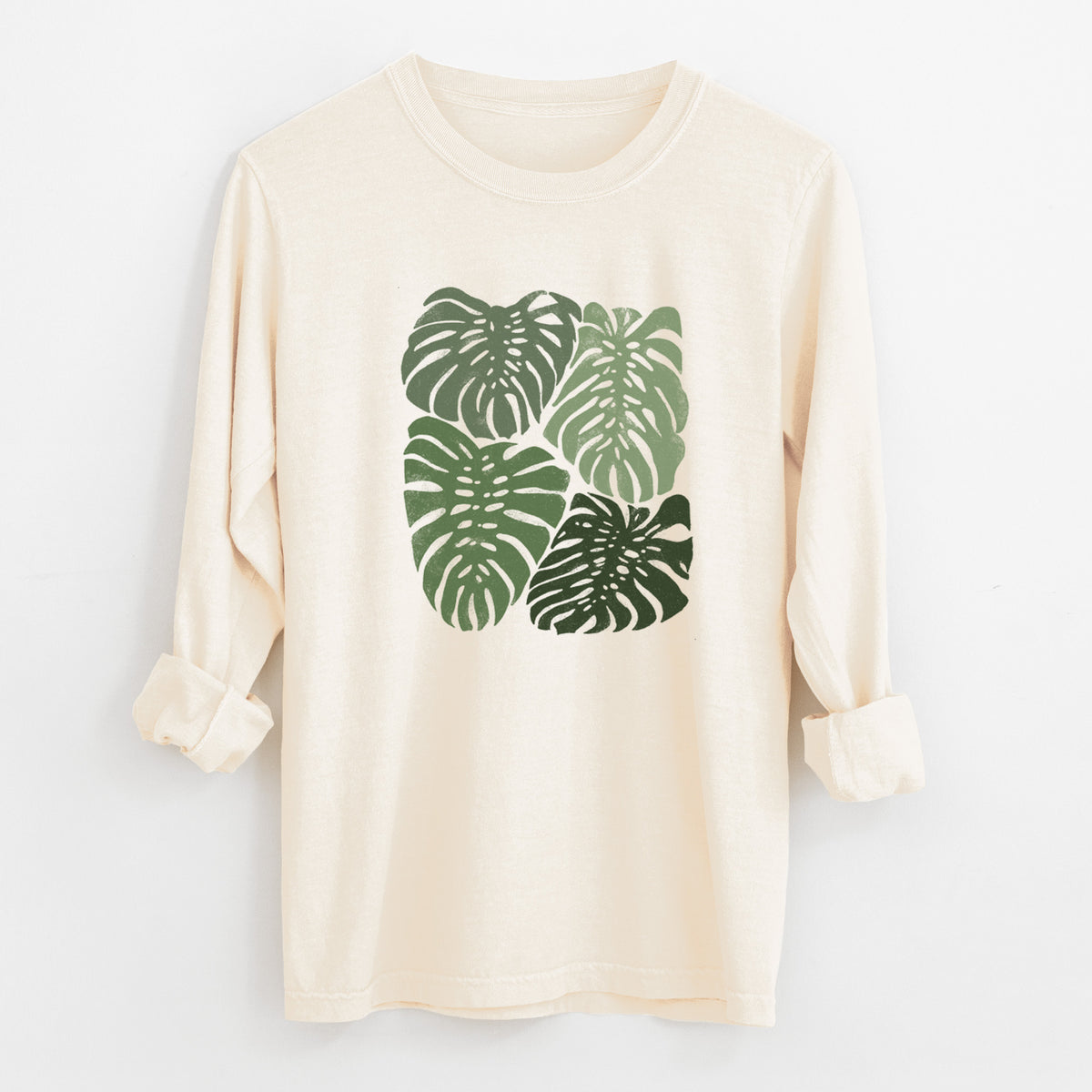 Ivory Comfort Colors long sleeve shirt laid flat with the cuffs rolled, printed with a full-color illustration of large green Monstera leaves filling a square.