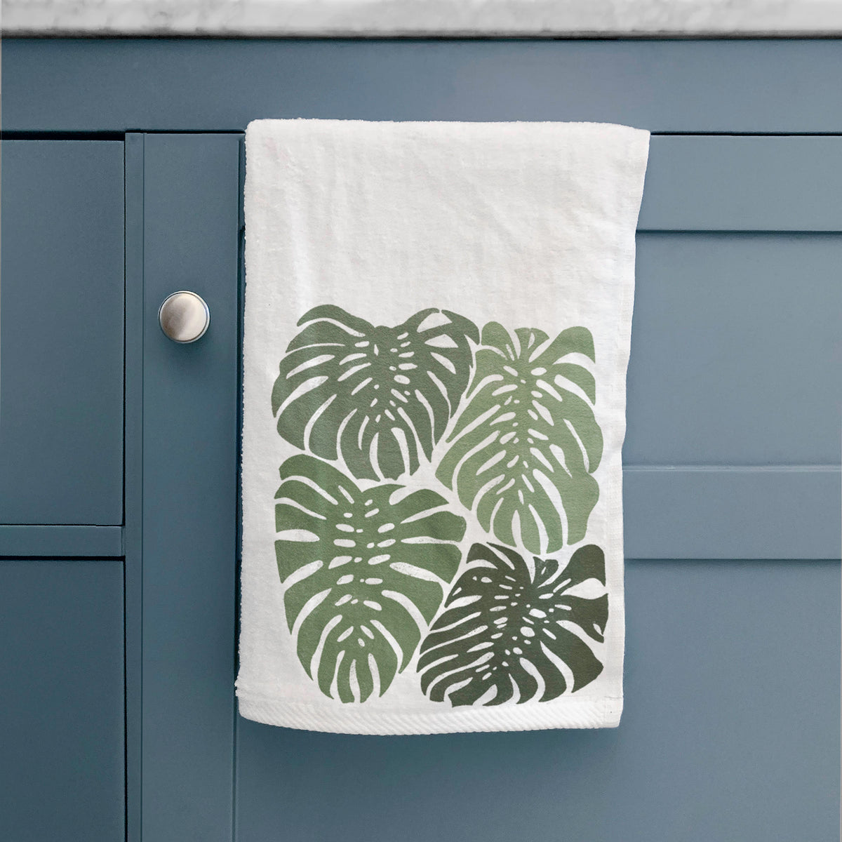 White hand towel printed with a full-color illustration of large green Monstera leaves filling a square, hanging on a cabinet door.