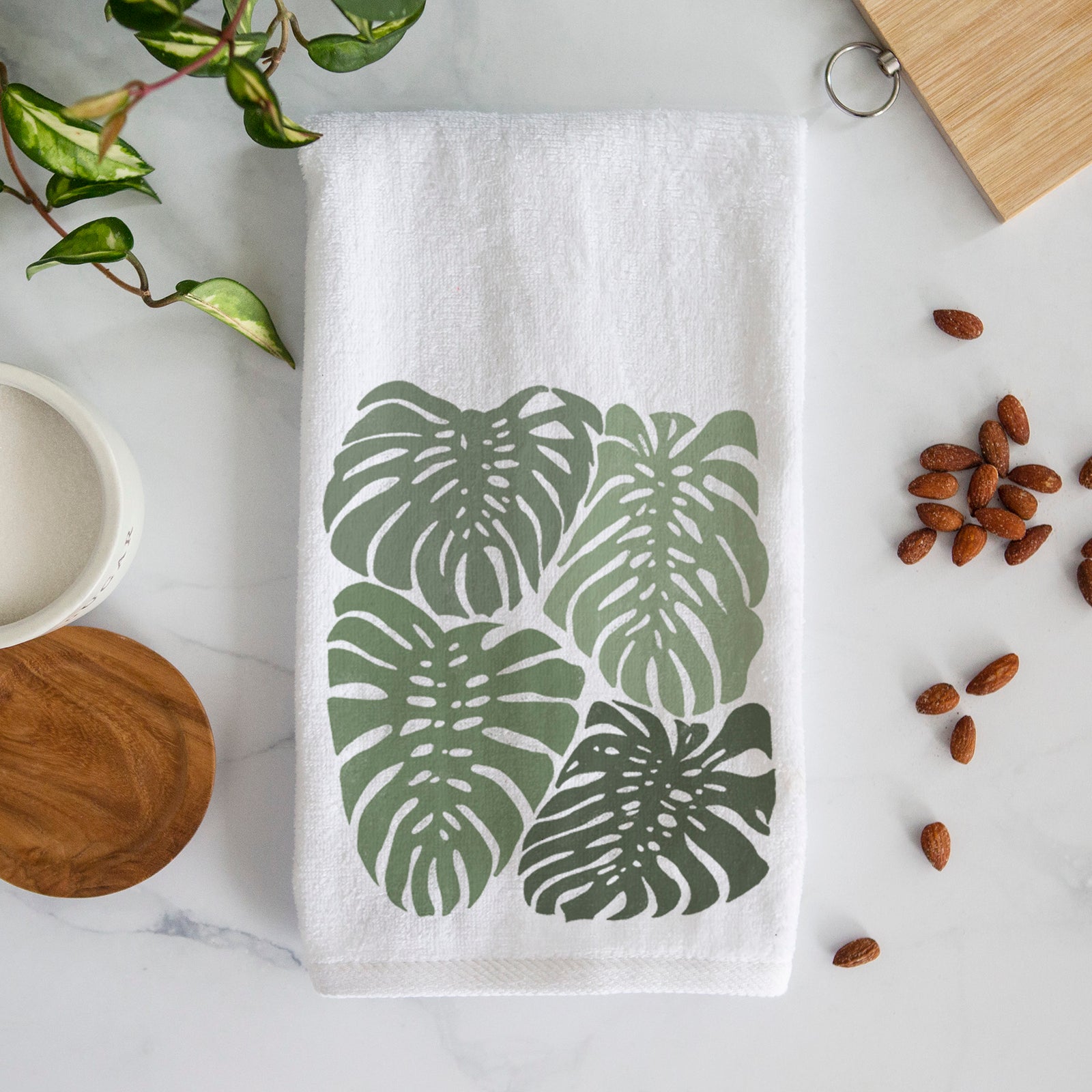 White hand towel printed with a full-color illustration of large green Monstera leaves filling a square.