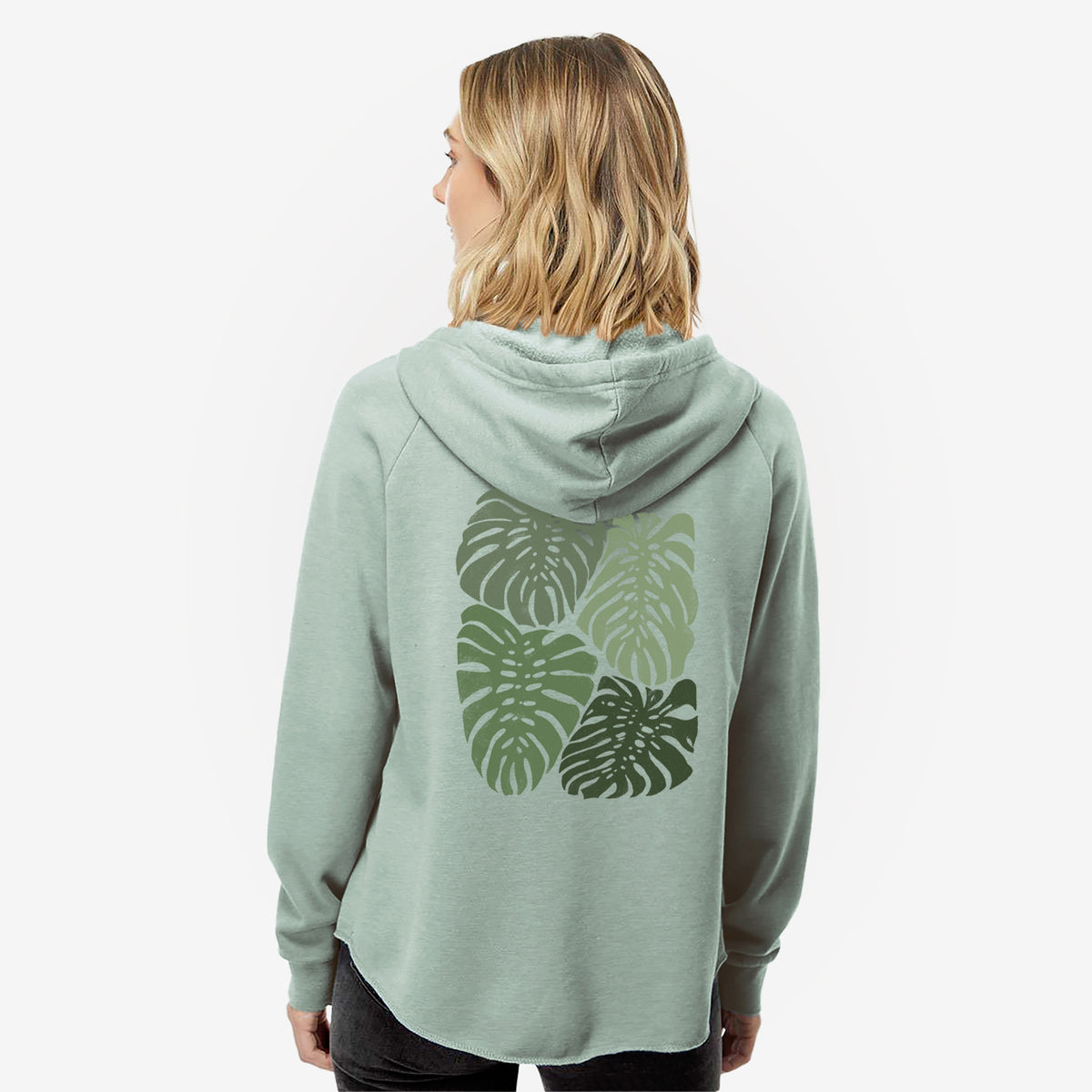 Woman wearing a sage green women's Cali Wave zip-up sweatshirt printed with a full-color illustration of large green Monstera leaves filling a square, seen from behind.