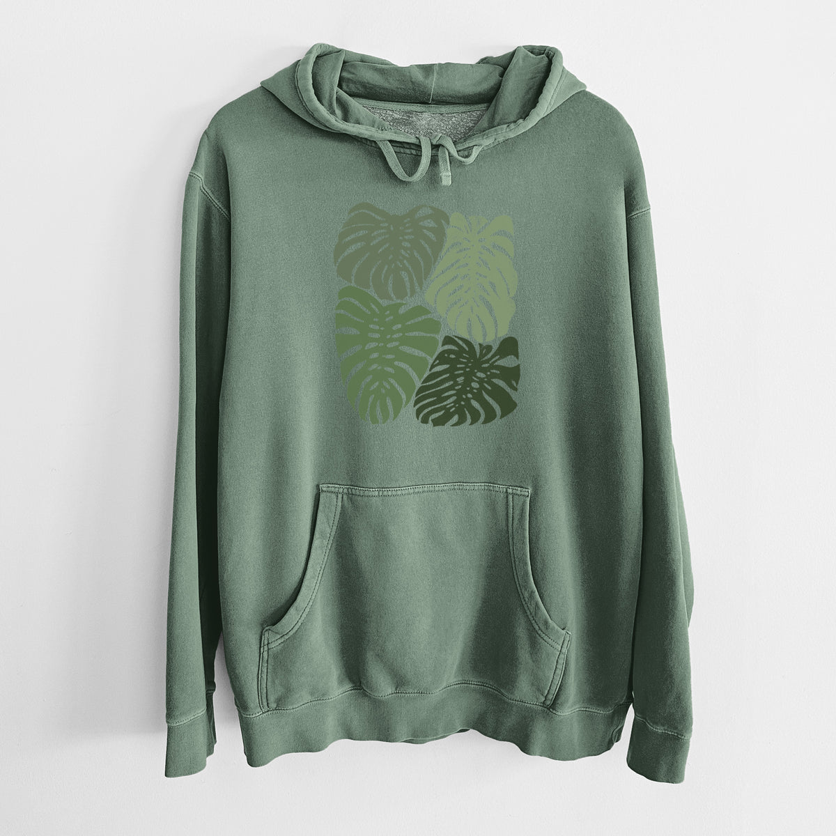 Alpine green pigment dyed hoodie laid flat, printed with a full-color illustration of large green Monstera leaves filling a square.