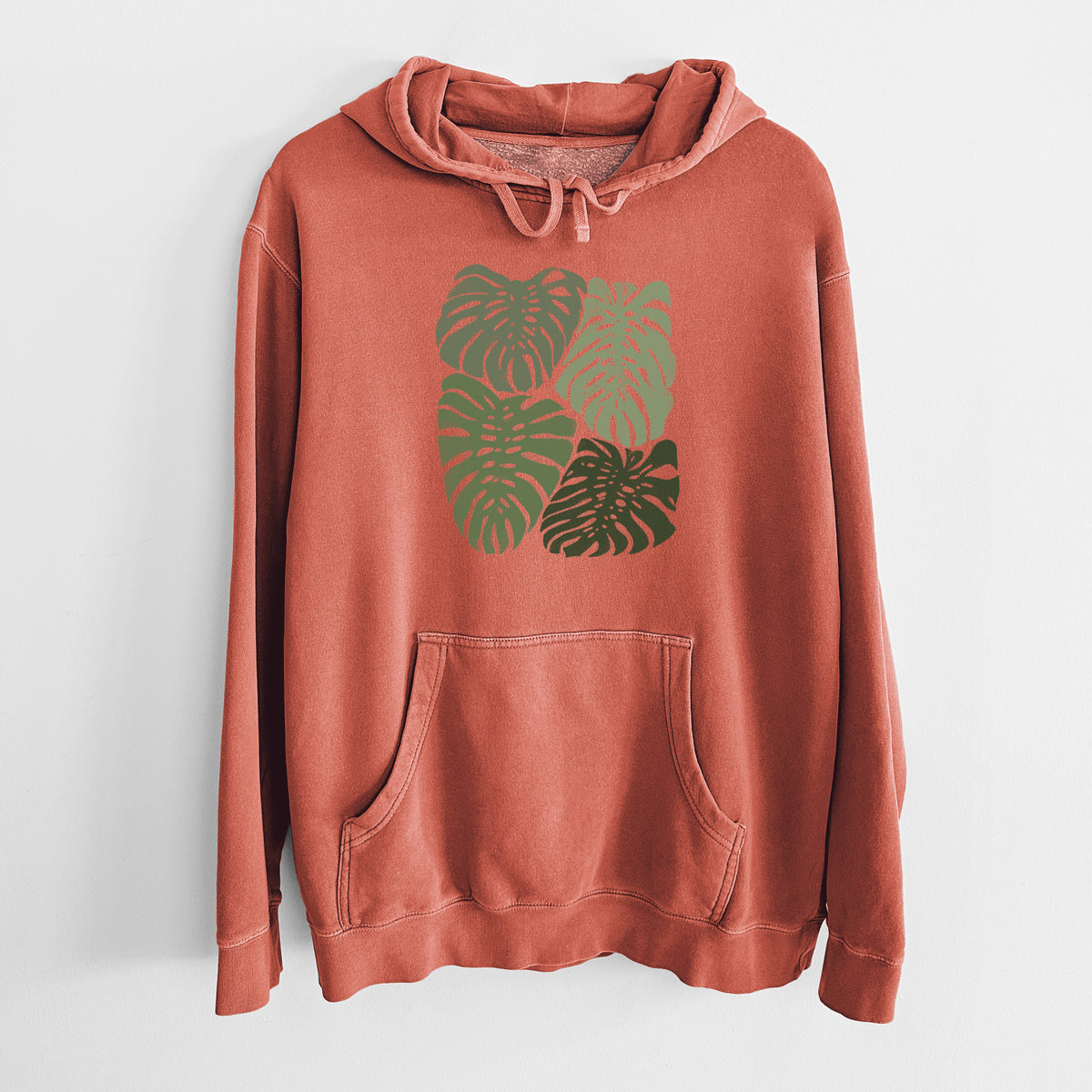 Amber orange pigment dyed hoodie laid flat, printed with a full-color illustration of large green Monstera leaves filling a square.